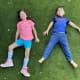 Two smiling children, a girl with cerebral palsy wearing ankle foot orthotics and a boy, lie in the grass together.
